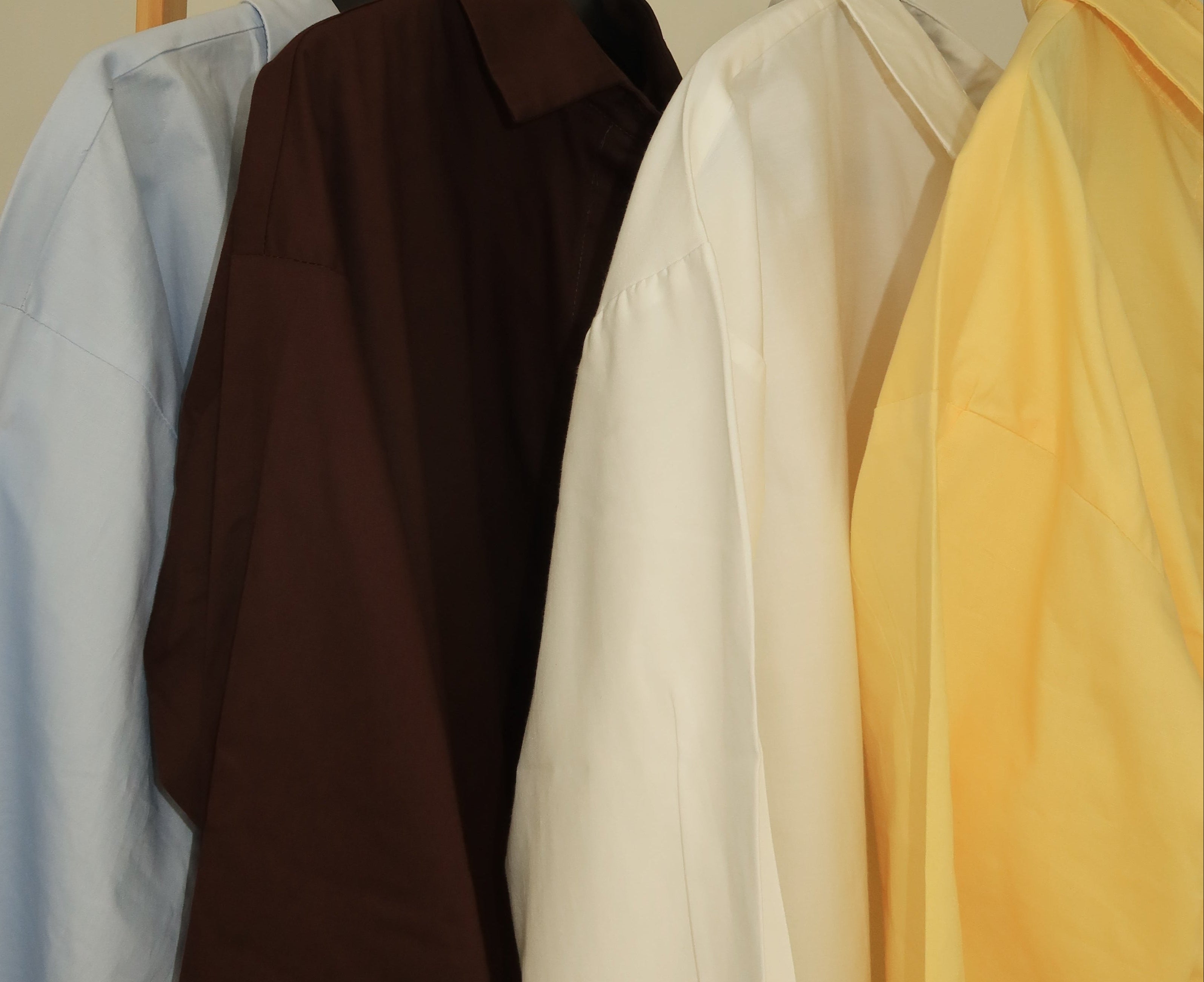Four long-sleeve button-up shirts in light blue, dark brown, white, and pale yellow hanging on black hangers from a wooden rack against a plain beige wall.